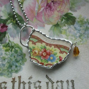 Könnte beinhalten: Halskette mit silberfarbener Kette und Anhänger aus einem Stück einer blumigen Teetasse. Der Anhänger hat ein farbenfrohes Blumenmuster mit roten, blauen und gelben Blumen. Eine bernsteinfarbene Perle hängt am Anhänger.
