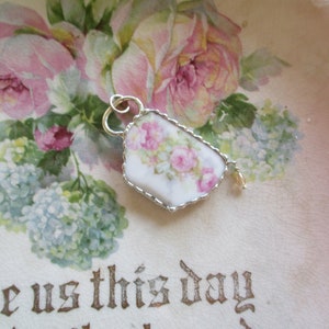 May include: A pendant crafted from a shard of white porcelain featuring a pink rose floral pattern, encased in a scalloped silver-toned metal frame with a small attached loop and a single dangling clear bead accent for jewelry wear.