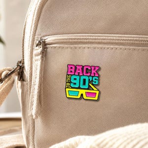 May include: A beige canvas backpack featuring a colorful enamel pin attached to the front pocket. The pin displays the text BACK TO THE 90'S in bright pink and cyan letters, positioned above a pair of yellow, cyan, and pink 3D glasses.