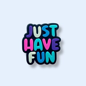 Op de afbeelding: Een kleurrijke rubberen charme met de tekst "JUST HAVE FUN" in een speels lettertype. De letters zijn in verschillende kleuren, waaronder roze, blauw, groen en paars.