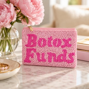 May include: A rectangular zippered pouch covered in light pink seed beads with the text "Botox Funds" embroidered in dark pink beads across the front. The pouch features a gold-toned metal zipper pull and a structured, flat-bottomed design.