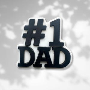 Może przedstawiać: Czarno-biały znak #1 Dad. Znak wykonany jest z grubego, białego materiału z czarnymi literami i cyframi.