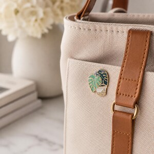 May include: A beige canvas tote bag with brown faux leather straps and a gold-toned metal pin featuring a potted monstera plant with light green and dark green leaves attached to the front pocket of the bag.
