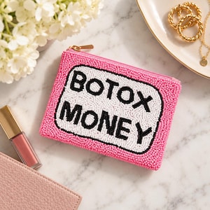 May include: A rectangular zippered pouch covered in pink beads with a central white beaded panel featuring the black beaded text BOTOX MONEY. The pouch has a gold-toned zipper pull and is constructed with a dense, textured beaded surface.