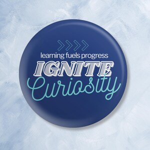 Può includere: Un bottone rotondo blu con il testo "learning fuels progress" e "IGNITE Curiosity" in lettere bianche e blu.