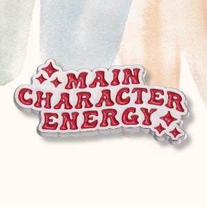 Puede incluir: Pin de esmalte blanco con letras rojas que dice "Main Character Energy" con dos pequeñas estrellas plateadas a cada lado.