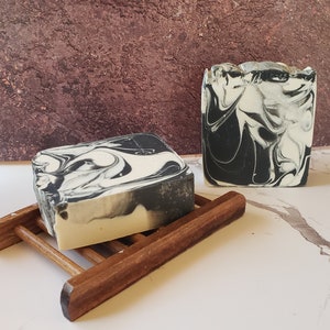 May include: Two bars of handmade soap with a black and white marbled design. The soap is sitting on a wooden soap dish.