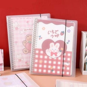 Op de afbeelding: Twee spiraalgebonden notitieboeken met doorschijnende covers en zwarte elastische sluitingen. Het voorste notitieboek toont een wit konijn in een hart, een rood-wit ruitpatroon en Japanse tekst. Het achterste notitieboek heeft een roze ruitpatroon.