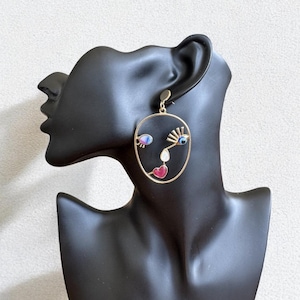 Könnte beinhalten: Goldfarbener Ohrring in Gesichtsform mit farbenfrohen Akzenten. Der Ohrring zeigt eine stilisierte Gesichtskontur mit einem blauen Auge, einem herzförmigen Mund in Rosa und einer weißen Nase. Der Ohrring wird auf einem schwarzen Schaufensterkopf präsentiert.