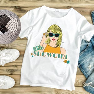 May include: White t-shirt featuring a cartoon illustration of a woman wearing sunglasses. The design includes the text "little SHOWGIRL" in teal and gold, with star accents. The woman has blonde hair and is wearing an orange top.