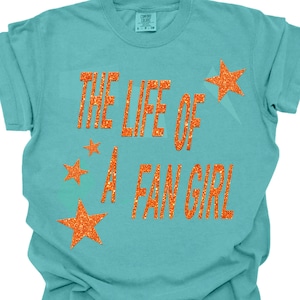 May include: A turquoise t-shirt with the text "THE LIFE OF A FAN GIRL" in orange glitter lettering. Orange glitter star graphics are scattered around the text, creating a fun and eye-catching design.