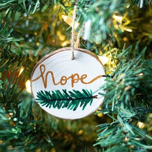 Pode incluir: Um enfeite de Natal de madeira com a palavra "Hope" pintada em letras douradas e um ramo de pinheiro verde pintado na parte inferior. O enfeite está pendurado em uma árvore de Natal verde.