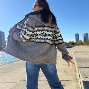 May include: A gray hooded zip-up sweater with a black and white snowflake pattern on the back. The sweater is worn with blue jeans. The background features a clear blue sky and a body of water.