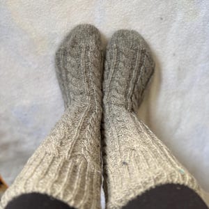 Pode incluir: Um par de meias de tricô cinza com cabo e textura canelada. As meias são usadas nas pernas de uma pessoa e puxadas até os joelhos.