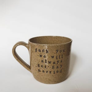 May include: A speckled brown ceramic mug with a curved handle. The mug features black text that reads "fuck you we will always get gay married."