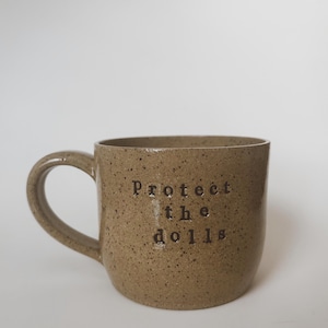 Puede incluir: Taza de cerámica moteada de color beige con asa curvada. La taza presenta el texto "Protect the dolls" en una fuente oscura sin serifa. La taza tiene un acabado brillante y un diseño sencillo y minimalista.