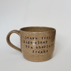 Puede incluir: Una taza de cerámica moteada de color marrón claro con un asa curva. La taza presenta un texto negro que dice "leave trans kids alone you absolute freaks". La taza está sobre un fondo blanco.