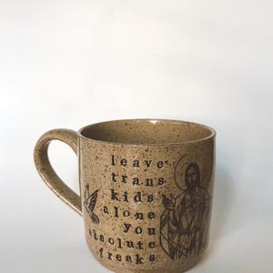 May include: A speckled beige ceramic mug with a handle. The mug features the text "leave trans kids alone you absolute freaks" in black, along with a bird and a religious figure illustration.