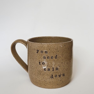 Op de afbeelding: Een gespikkelde bruine keramische mok met een gebogen handvat. De mok heeft de zwarte tekst "you need to calm down" verticaal afgedrukt. De mok is neutraal van kleur en heeft een eenvoudig, minimalistisch ontwerp.