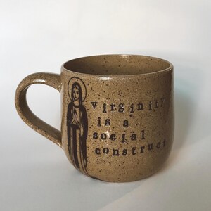 May include: A speckled brown ceramic mug with a handle. The mug features a dark brown illustration of a saint and the text "Virginity is a social construct" in a dark brown font.