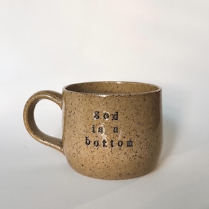 May include: A speckled tan ceramic mug with a curved handle. The mug has the words "god is a bottom" printed in black, stacked vertically. The mug is set against a white background.