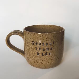 Puede incluir: Una taza de cerámica marrón moteada con un asa curvada. La taza presenta el texto "protect trans kids" en letras negras. La taza está colocada sobre un fondo blanco liso.