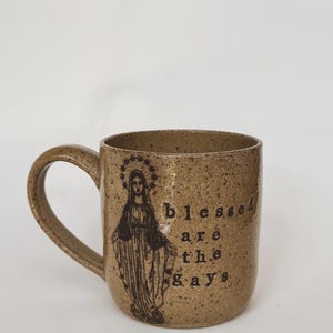 Puede incluir: Una taza de cerámica marrón moteada con asa. La taza presenta una ilustración de una figura religiosa y las palabras "blessed are the gays" en letras negras. La taza tiene un acabado brillante.