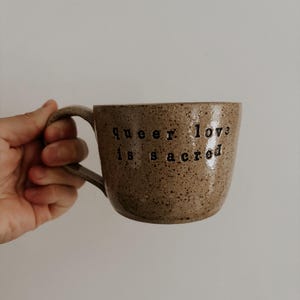 Puede incluir: Una taza de cerámica marrón moteada con un asa negra. La taza tiene el texto "Queer love is sacred" impreso en el lateral.