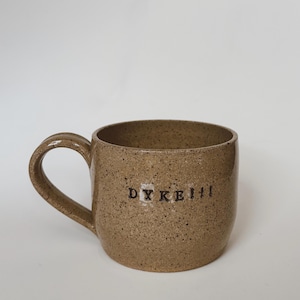 Puede incluir: Una taza de cerámica moteada de color beige con un asa curvada. La taza presenta la palabra "DYKE!!!" en letras negras. La taza es de color neutro y tiene un diseño sencillo y funcional.