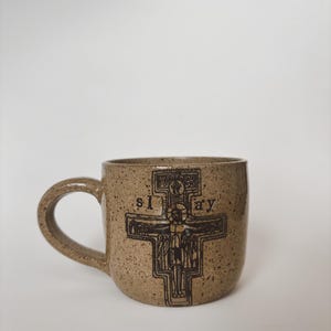 Può includere: Una tazza in ceramica beige macchiettata con manico. La tazza presenta un'illustrazione marrone scuro di un crocifisso con la parola "slay" stampata sul davanti. La tazza è posta su uno sfondo bianco.