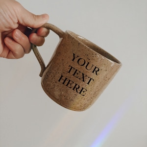 Puede incluir: Una taza de cerámica marrón moteada con asa. La taza tiene el texto "YOUR TEXT HERE" impreso en el lateral.