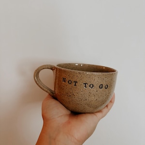 Puede incluir: Una taza de cerámica marrón moteada con forma redondeada y asa. La taza tiene el texto "HOT TO GO" impreso en el lateral.