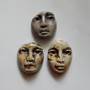 May include: Three ceramic face sculptures with a glossy finish. The top piece is a cool-toned grey, while the two bottom pieces feature cream and brown mottled glazes. Each sculpture depicts a human face with defined eyes, nose, and lips.