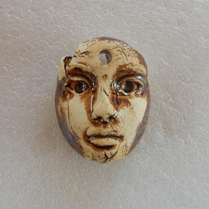 May include: A ceramic face pendant with a hole at the top for hanging. The face is a light beige color with brown accents around the eyes, nose, and mouth. The pendant has a distressed look, with a chipped edge on the left side.