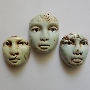 May include: Three ceramic face sculptures in a pale blue glaze with dark brown crackle detailing. Each piece features distinct facial expressions, with the central face showing spiral forehead carvings and the others displaying weathered, textured surfaces.