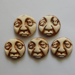 May include: Five circular ceramic cabochons, each featuring a molded human face with a cream-colored glaze and dark brown accents in the recessed areas of the eyes, nose, and mouth, arranged in a group of two on top and three below.