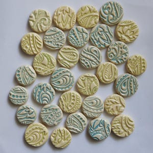 May include: A collection of small, round ceramic beads with embossed floral and abstract patterns. The beads are primarily cream-colored, with accents of light green and teal. The designs vary, creating a textured and artistic appearance.