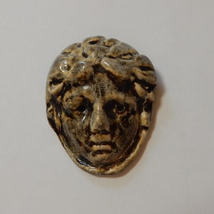 May include: A ceramic brooch shaped like a human face with a glossy, mottled beige and dark brown glaze. The sculpted features include defined eyes, a nose, and lips, with textured, wavy hair framing the forehead and sides of the face.