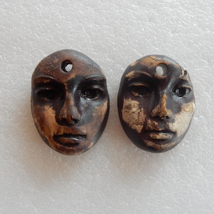 May include: Two oval-shaped pendants featuring sculpted human faces in a dark brown finish with distressed beige highlights. Each pendant has a circular hole at the top for suspension, showcasing detailed facial features including eyes, nose, and lips.