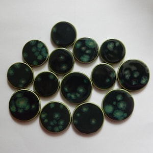 May include: A collection of thirteen circular ceramic buttons featuring a glossy dark black glaze with scattered, soft-edged teal and green crystalline patterns across the surface of each individual piece, arranged in a loose cluster.