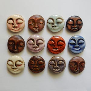 May include: Twelve circular ceramic cabochons arranged in a grid, each featuring a sculpted human face with closed eyes and a serene expression. The pieces display a variety of glazed finishes in shades of cream, brown, blue, orange, and purple.