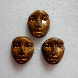 May include: Three oval-shaped ceramic face sculptures with a glossy, mottled brown glaze. Each piece features molded facial details including eyes, a nose, and lips, with a smooth, rounded surface and dark brown highlights across the features.