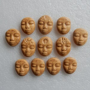 May include: A collection of eleven small, tan-colored ceramic face beads with closed eyes. Some beads feature etched designs like a spiral, a crescent moon, a peace symbol, and the letters "TO VE". The beads have a speckled texture.