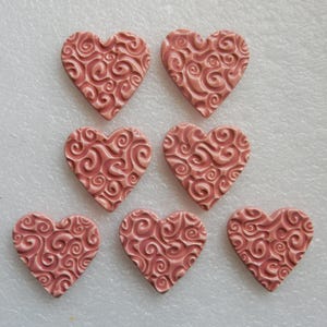 May include: Seven pink heart-shaped decorative pieces with a swirling, embossed pattern. The hearts are arranged on a white surface. These decorative hearts could be used for crafts or embellishments.