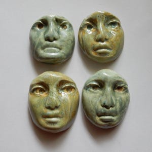 May include: Four ceramic face sculptures with a distressed, aged appearance. The faces are oval-shaped with detailed features, including eyes, noses, and mouths. The color palette includes shades of green, yellow, and brown, giving them an earthy aesthetic.