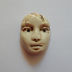 May include: A sculpted ceramic face cabochon in an off-white cream color with dark brown crackle detailing across the forehead, eyes, and cheeks. The facial features are molded with depth, including defined eyes, a nose, and lips.