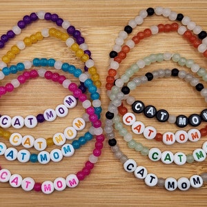 May include: Five colorful beaded bracelets with the words "CAT MOM" spelled out in white letters. The bracelets are made with translucent beads in shades of purple, yellow, turquoise, pink, and white.