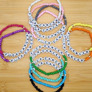 May include: A collection of beaded bracelets in various colors, including purple, pink, green, blue, and orange. Each bracelet features white letter beads spelling "FUCK CANCER". The bracelets are arranged on a wooden surface.