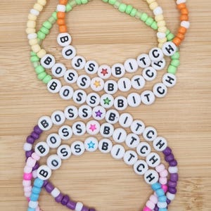 May include: Stack of beaded bracelets in orange, yellow, green, purple, pink, and blue. The bracelets spell out "BOSS BITCH" with white letter beads and star accents. The bracelets are on a light brown wooden surface.