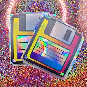 Puede incluir: Dos pegatinas holográficas iridiscentes de disquetes. Las pegatinas son plateadas con un efecto arcoíris y tienen un esquema de colores amarillo, naranja, rojo, azul y morado.